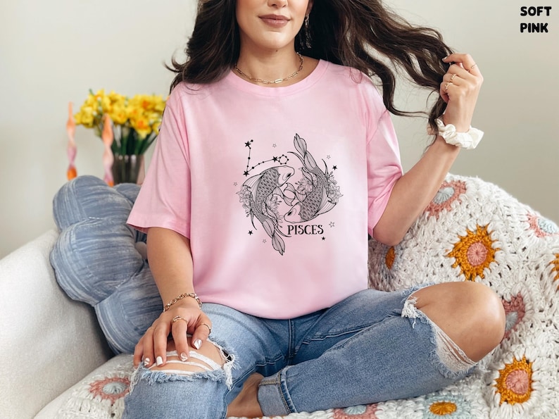 May include: A soft pink t-shirt featuring a black and white Pisces design. The graphic includes two fish, stars, and the word "PISCES". The shirt is made of a comfortable material.