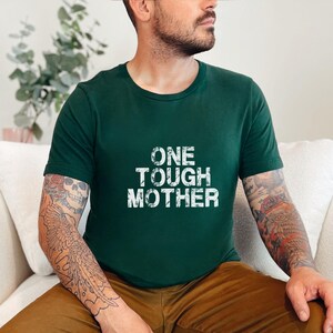 May include: A person wearing a forest green t-shirt with the text "ONE TOUGH MOTHER" printed in white. The person has tattoos on their arms.
