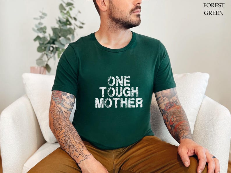 May include: A person wearing a forest green t-shirt with the text "ONE TOUGH MOTHER" printed in white. The person has tattoos on their arms.