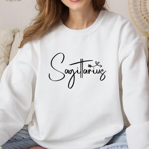 K&ouml;nnte beinhalten: Wei&szlig;er Langarm-Pullover mit dem Wort "Sch&uuml;tze" in schwarzer Kursivschrift mit einem kleinen Pfeil- und Blattdesign.