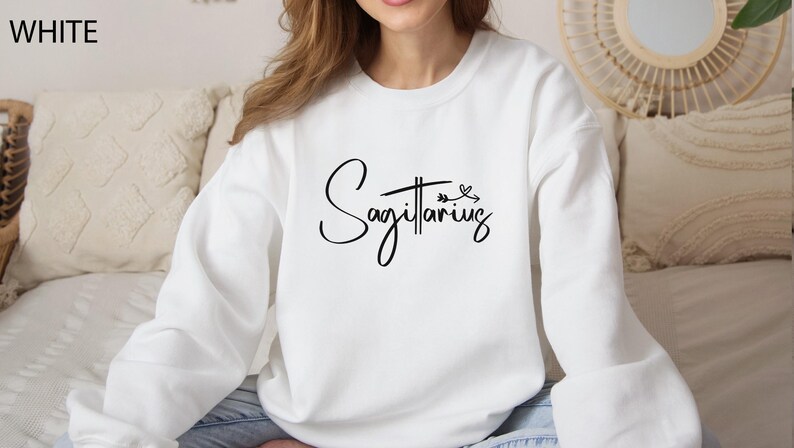 K&ouml;nnte beinhalten: Wei&szlig;er Langarm-Pullover mit dem Wort "Sch&uuml;tze" in schwarzer Kursivschrift mit einem kleinen Pfeil- und Blattdesign.
