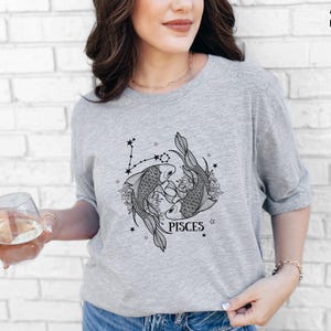 May include: Athletic heather gray t-shirt featuring a black and white Pisces zodiac design. The graphic includes two fish, floral elements, and the word "PISCES". The shirt is made of a soft material.