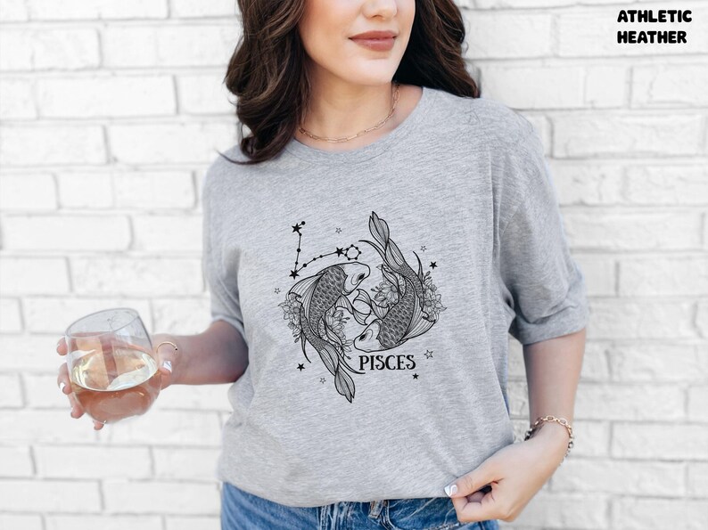 May include: Athletic heather gray t-shirt featuring a black and white Pisces zodiac design. The graphic includes two fish, floral elements, and the word "PISCES". The shirt is made of a soft material.