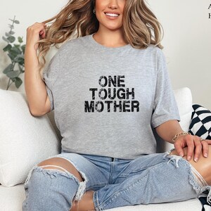 May include: A gray t-shirt with the text "ONE TOUGH MOTHER" printed in black. The shirt is being worn by a person sitting on a white couch.