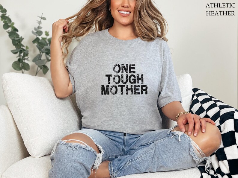 May include: A gray t-shirt with the text "ONE TOUGH MOTHER" printed in black. The shirt is being worn by a person sitting on a white couch.