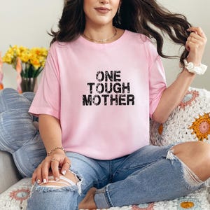 May include: A woman wearing a pink t-shirt with the text "ONE TOUGH MOTHER" in black. She is sitting on a white crocheted blanket with a blue pillow behind her.