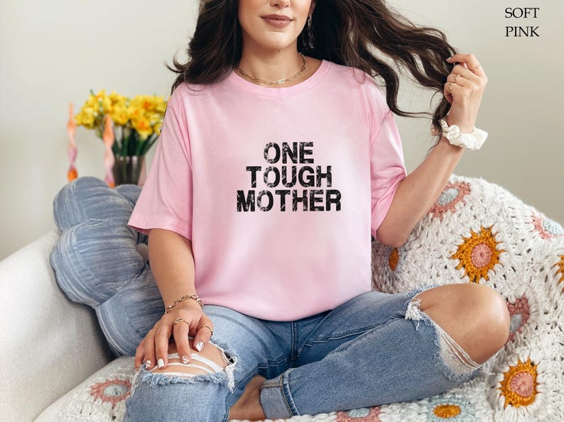May include: A woman wearing a pink t-shirt with the text "ONE TOUGH MOTHER" in black. She is sitting on a white crocheted blanket with a blue pillow behind her.