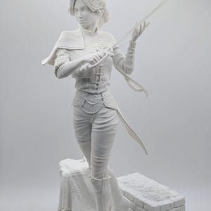 Peut inclure: Sculpture blanche d'une personne en tenue détaillée, tenant une épée. La figurine se tient sur une plateforme en planches de bois et en briques. La sculpture a une cape et est dans une pose dynamique.