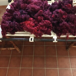 May include: A pile of deep purple and red wool fibers on a white frame with a black metal bar. The fibers are loose and fluffy, and they are arranged in a random pattern.