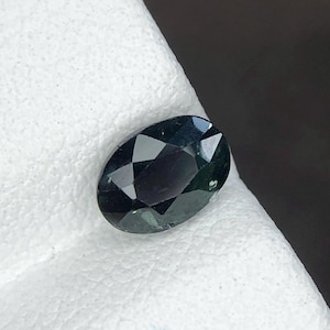 Puede incluir: Una piedra preciosa de corte ovalado con un tono verde oscuro profundo y una superficie facetada que refleja la luz. La piedra presenta un acabado pulido con bordes geométricos definidos, mostrando una calidad translúcida y variada.