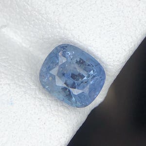 Puede incluir: Una piedra preciosa de color azul claro con talla cojín y superficie facetada. La piedra muestra una calidad translúcida con inclusiones internas visibles y un acabado pulido que refleja la luz a través de sus múltiples facetas.