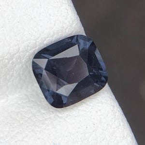 Puede incluir: Una piedra preciosa de talla cojín con un tono azul grisáceo profundo y oscuro, con una superficie facetada que refleja la luz. La piedra presenta una forma cuadrada con esquinas redondeadas y un aspecto pulido y translúcido.