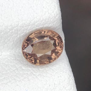 Puede incluir: Una piedra preciosa de corte ovalado con un tono cálido marrón claro y melocotón. La piedra presenta una superficie facetada que refleja la luz, creando una apariencia clara y multifacética con claridad interna visible y un acabado pulido.