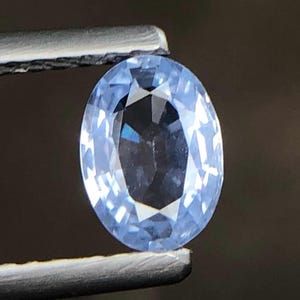 Puede incluir: Una piedra preciosa de color azul claro con corte ovalado y superficie facetada que refleja la luz. La piedra está sujeta por unas pinzas metálicas, mostrando su claridad transparente y su perfil de color frío, creando un aspecto brillante.
