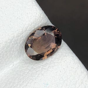 Puede incluir: Una piedra preciosa de corte ovalado con un tono marrón translúcido y una superficie facetada. La piedra presenta un acabado pulido con múltiples facetas geométricas que reflejan la luz, creando una apariencia clara y con múltiples tonos.