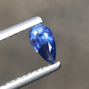 Puede incluir: Una piedra preciosa azul en forma de pera con una superficie facetada sujeta por unas pinzas de metal. La piedra muestra un color azul real profundo con reflejos de luz internos y bordes afilados, exhibiendo una calidad transparente.
