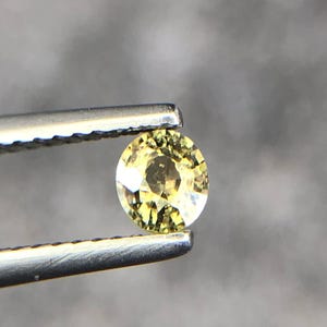 Puede incluir: Una piedra preciosa amarilla redonda y facetada sujeta por unas pinzas de metal plateado. La piedra muestra un tono dorado brillante y translúcido con múltiples reflejos internos y facetas geométricas nítidas en su superficie.