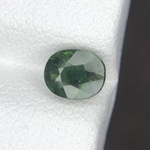 Puede incluir: Una piedra preciosa de corte ovalado con un tono verde bosque profundo y una superficie multifacetada. La piedra muestra claridad interna y reflexión de la luz a través de sus facetas pulidas y simétricas, exhibiendo un perfil de color rico.