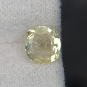 Puede incluir: Una piedra preciosa amarilla de talla cojín con una superficie facetada que refleja la luz. La piedra muestra un tono amarillo limón pálido translúcido y está colocada sobre una superficie de tela suave, texturizada y de color blanco roto.