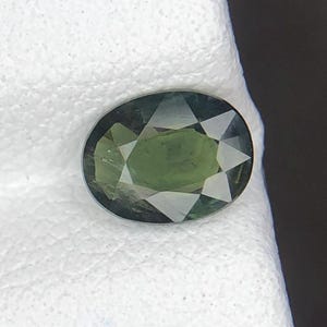 Puede incluir: Una piedra preciosa de corte ovalado con un tono verde oliva profundo y una superficie facetada que crea un patrón de reflejos de luz en forma de estrella en la parte superior. La piedra muestra tonos variados de verde oscuro y claro.