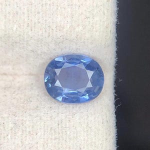 Puede incluir: Una piedra preciosa de zafiro azul con corte ovalado y superficie facetada que refleja la luz. La gema muestra un tono azul aciano vibrante con una claridad interna nítida y una disposición simétrica de facetas triangulares y en forma de cometa.