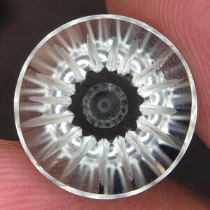 Könnte beinhalten: Ein runder Knopf aus klarem Glas mit einem sternförmigen Muster aus geätzten Strahlen, die von einem zentralen schwarzen, gezackten Kern ausgehen. Das Design erzeugt einen geometrischen Strahlenkranzeffekt innerhalb des transparenten Materials.