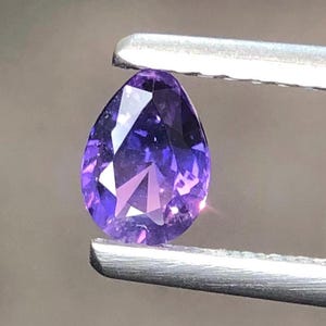 Puede incluir: Una piedra preciosa morada con facetas en forma de pera, sostenida por unas pinzas de metal plateado. La piedra muestra un tono violeta vibrante con múltiples facetas triangulares y en forma de diamante que reflejan la luz.