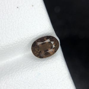 Puede incluir: Una piedra preciosa de corte ovalado con un tono marrón translúcido y una superficie facetada. La piedra muestra un patrón claro de múltiples facetas en la parte superior, reflejando la luz a través de su cuerpo de tonos tierra oscuros.
