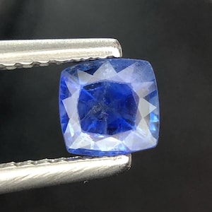 Puede incluir: Una piedra preciosa de zafiro azul con corte cojín y superficie facetada, sostenida por unas pinzas de metal plateado. La gema muestra un color azul profundo y translúcido con inclusiones internas visibles y facetas geométricas nítidas.