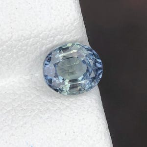 May include: An oval-cut gemstone with a light blue and pale green colour palette. The stone features a faceted surface that reflects light, creating a sparkling appearance with visible internal clarity and a mix of cool-toned hues throughout the gem.
