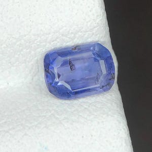 Puede incluir: Una piedra preciosa azul rectangular facetada con un corte escalonado. La piedra muestra un tono azul bígaro translúcido con inclusiones oscuras visibles y características minerales naturales en toda la estructura cristalina.