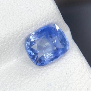 May include: A cushion-cut blue sapphire gemstone with a faceted surface. The stone displays a vibrant cornflower blue hue with internal light reflections and visible clarity characteristics throughout the crystalline structure of the gem.