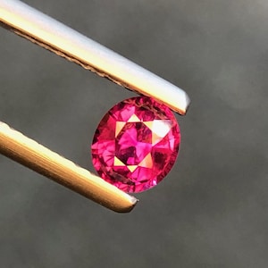 Puede incluir: Una gema de corte ovalado vibrante con un tono magenta rosado intenso y una superficie multifacetada, sostenida por unas pinzas metálicas. La piedra muestra reflejos de luz internos y un acabado brillante y pulido en toda su estructura.