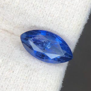 Puede incluir: Una piedra preciosa de corte marquesa con un color azul profundo y vibrante y una superficie facetada que refleja la luz. La piedra tiene una forma ovalada alargada y puntiaguda, presentada sobre una tela texturizada de color blanco roto.