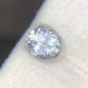 Puede incluir: Una piedra preciosa incolora de corte ovalado con múltiples facetas que reflejan la luz, creando un aspecto brillante y centelleante. La piedra es clara y transparente, mostrando un alto nivel de claridad y una superficie pulida y reflectante.
