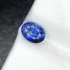 May include: A faceted oval-shaped blue sapphire gemstone with a brilliant cut, displaying deep royal blue tones and internal light reflections across its polished surface, showcasing a clear and transparent crystalline structure.