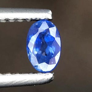Puede incluir: Una gema azul vibrante de corte ovalado con una superficie facetada sostenida por pinzas de metal. La piedra muestra un color azul profundo y saturado con reflejos de luz internos en sus facetas pulidas, con aspecto transparente.
