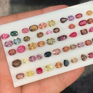 Puede incluir: Una bandeja de presentación blanca que contiene una colección de numerosas piedras preciosas sueltas de diversas formas y tamaños. Las piedras presentan un amplio espectro de colores, incluyendo rosa, amarillo, marrón, azul, rojo y morado, dispuestas en filas.