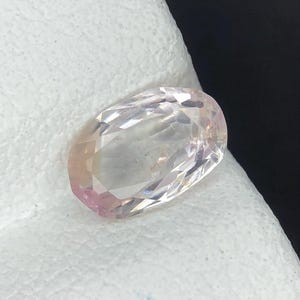 Puede incluir: Una piedra preciosa de corte ovalado con una superficie facetada, que muestra un tono rosa blanquecino pálido y translúcido. La piedra presenta una calidad clara y reflectante con múltiples facetas geométricas que captan la luz.