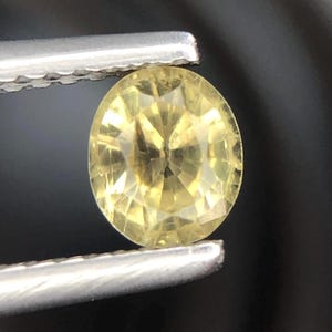 Puede incluir: Una piedra preciosa amarilla de forma ovalada con facetas y corte brillante, que muestra reflejos de luz internos y claridad. La piedra se sostiene entre las puntas metálicas de unas pinzas, exhibiendo su tono dorado translúcido y facetas geométricas.