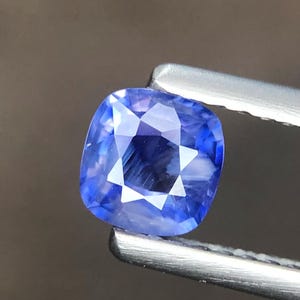 Puede incluir: Una piedra preciosa de zafiro azul con corte tipo cojín y superficie facetada, sostenida por unas pinzas de metal plateado. La gema muestra un color azul vibrante y translúcido con reflejos de luz internos y bordes geométricos definidos.