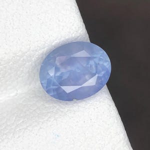 Puede incluir: Una piedra preciosa de corte ovalado en color azul bígaro claro con una superficie facetada. La piedra muestra una calidad translúcida con claridad interna y un acabado pulido, reflejando la luz a través de sus múltiples facetas geométricas.