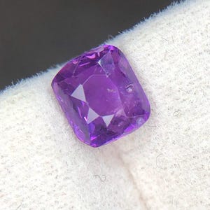 Puede incluir: Una piedra preciosa de corte cojín con un tono púrpura vibrante y una superficie facetada. La piedra muestra una calidad clara y transparente con bordes afilados y un acabado pulido, reflejando la luz a través de sus múltiples facetas.