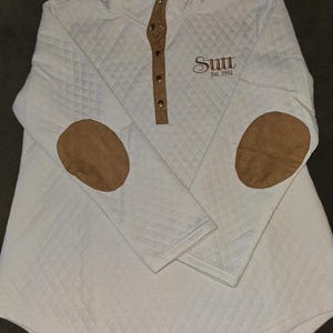 Puede incluir: Sudadera blanca acolchada con parches de ante marrón en los codos y una plaqueta de botones a presión. La sudadera tiene el texto "Sutt Est. 1992" bordado en la parte delantera.