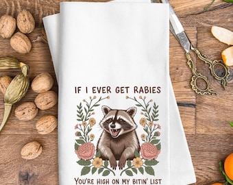 Könnte beinhalten: Weißes Küchentuch mit einer Illustration eines Waschbären, umgeben von Blumen. Der Text lautet: „IF I EVER GET RABIES YOU'RE HIGH ON MY BITIN' LIST.“