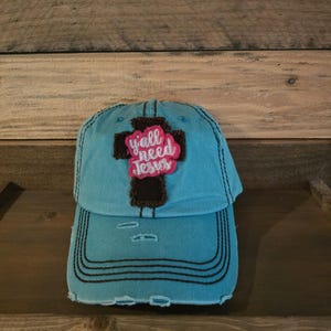 May include: A turquoise blue baseball cap with a distressed look and black stitching. The cap has a brown and black cross patch with the words "y'all need Jesus" in pink lettering.