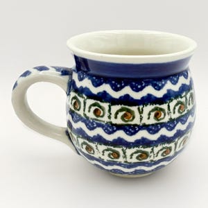 Puede incluir: Una taza de cerámica con interior blanco y borde azul. El exterior presenta un patrón repetitivo de ondas azules, detalles blancos y diseños florales verdes y marrones. El asa es de color gris claro.