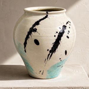 May include: A ceramic vase with a cream-colored base, adorned with bold black paint splatters and a splash of turquoise. The vase has a rounded body and a wide opening, suitable for displaying flowers or as a decorative piece.