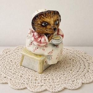 May include: A ceramic figurine of a hedgehog wearing a bonnet and dress, seated on a small stool. The hedgehog is brown with a white bonnet and a dress with red floral accents. It holds a small dish. The figurine sits on a cream-colored, crocheted doily.