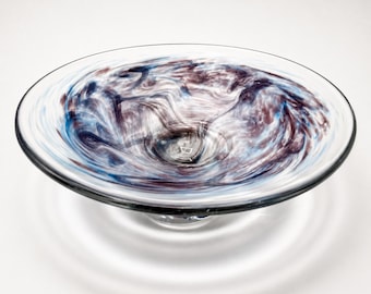 May include: A clear glass bowl with a swirling pattern of blue, white, and maroon. The bowl has a wide, shallow design and sits on a small, clear base. The colors blend together, creating a marbled effect.